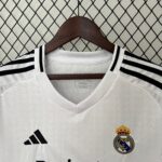 White Real Madrid soccer jersey on a wooden hanger, with the adidas logo and Real Madrid crest visible, set against a diamond-pattern chain-link fence.