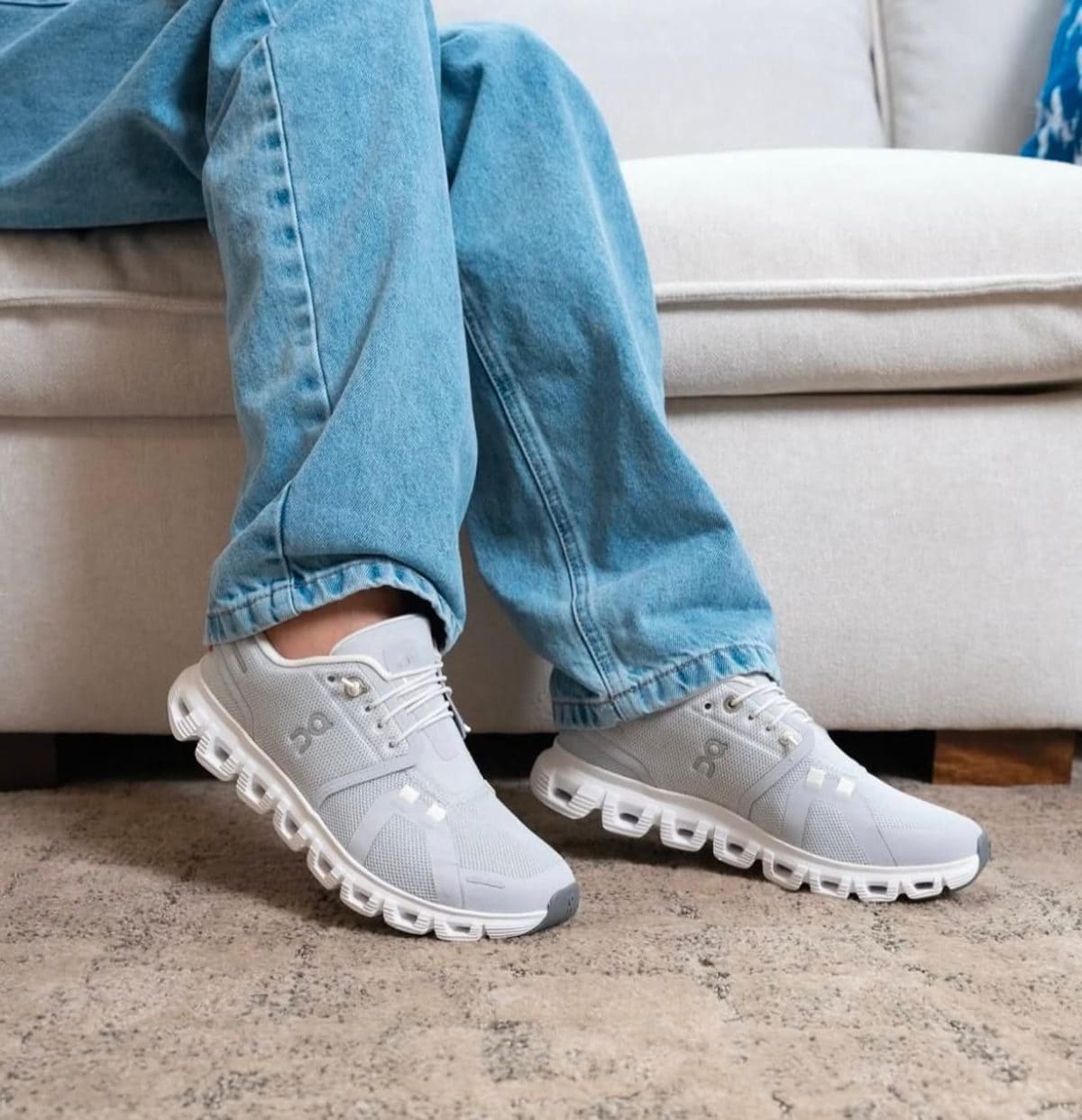 Person wearing light gray athletic sneakers and blue jeans, seated on a sofa, showing the side and sole of the shoes