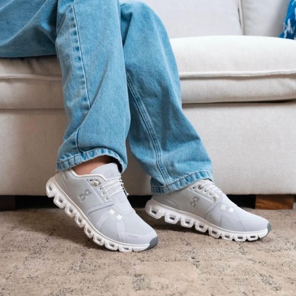 Person wearing light gray athletic sneakers and blue jeans, seated on a sofa, showing the side and sole of the shoes