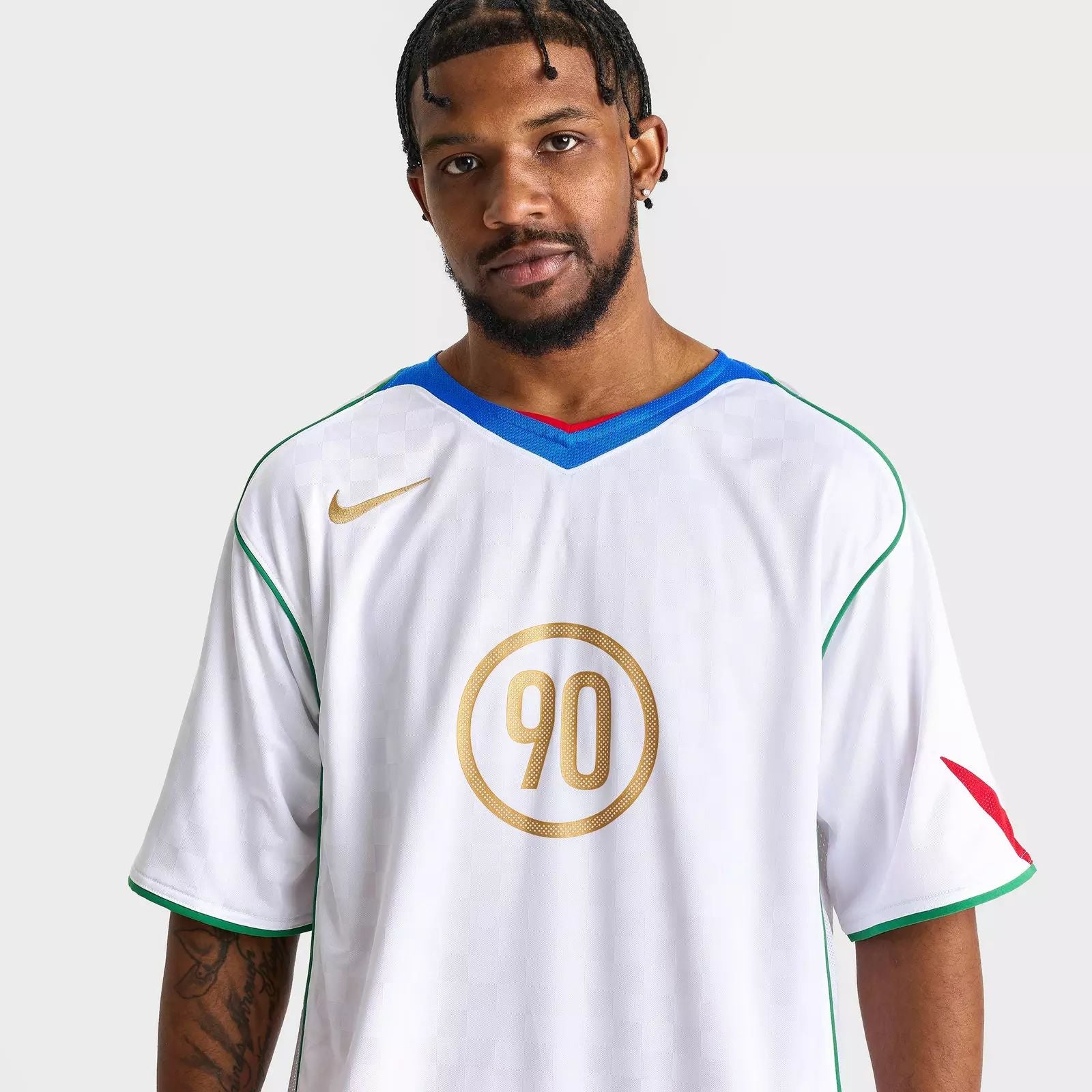 6558f050-9584-4b69-b3a8-23afdb374918 Portrait of a man with short dreadlocks and a beard, wearing a white sports jersey with blue trim and green piping, featuring a large gold circular 90 emblem on the chest against a light gray background.