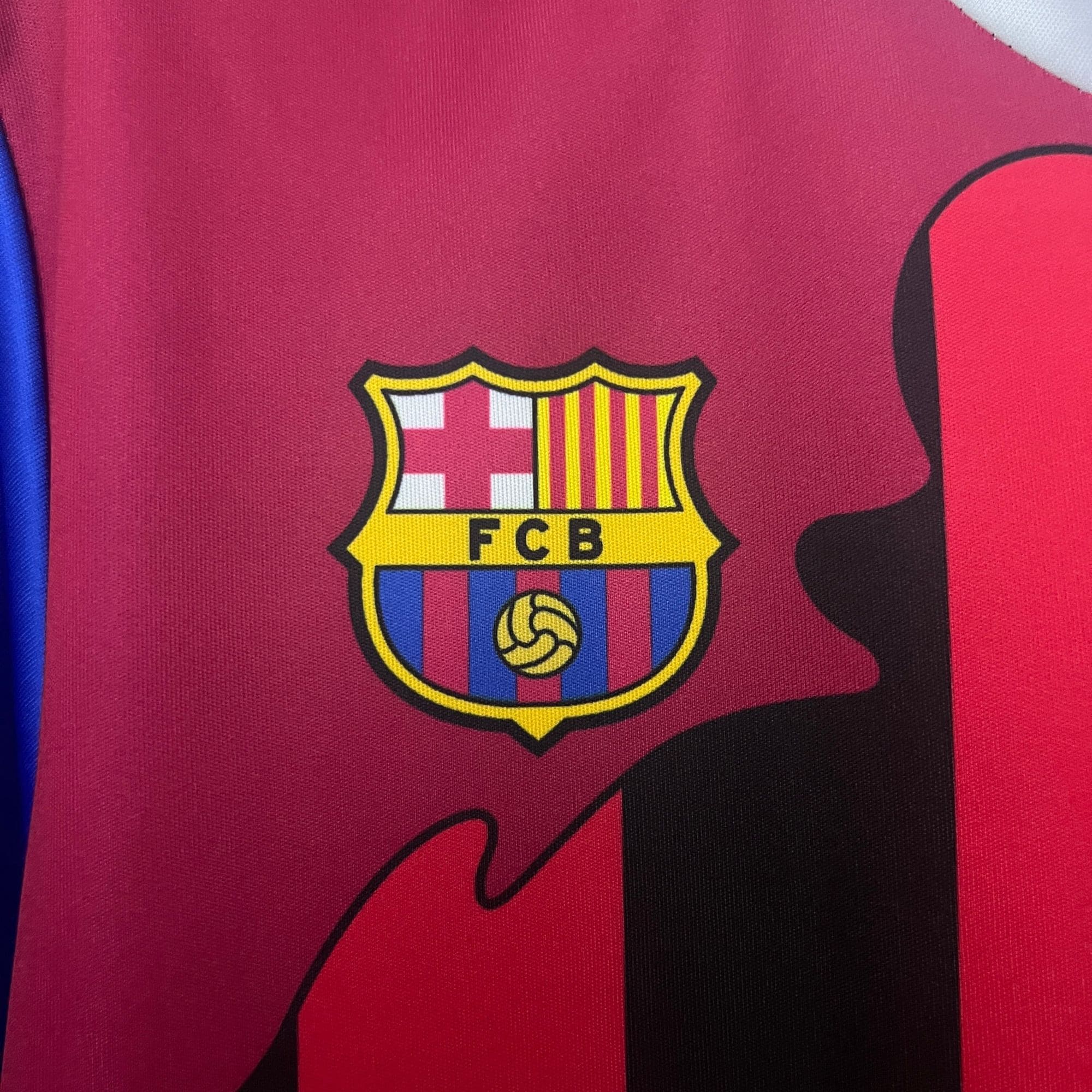 65bec125 | UAE SPORT STORE Close-up of a red FC Barcelona jersey showing part of the club crest and a blue stripe along the side.
