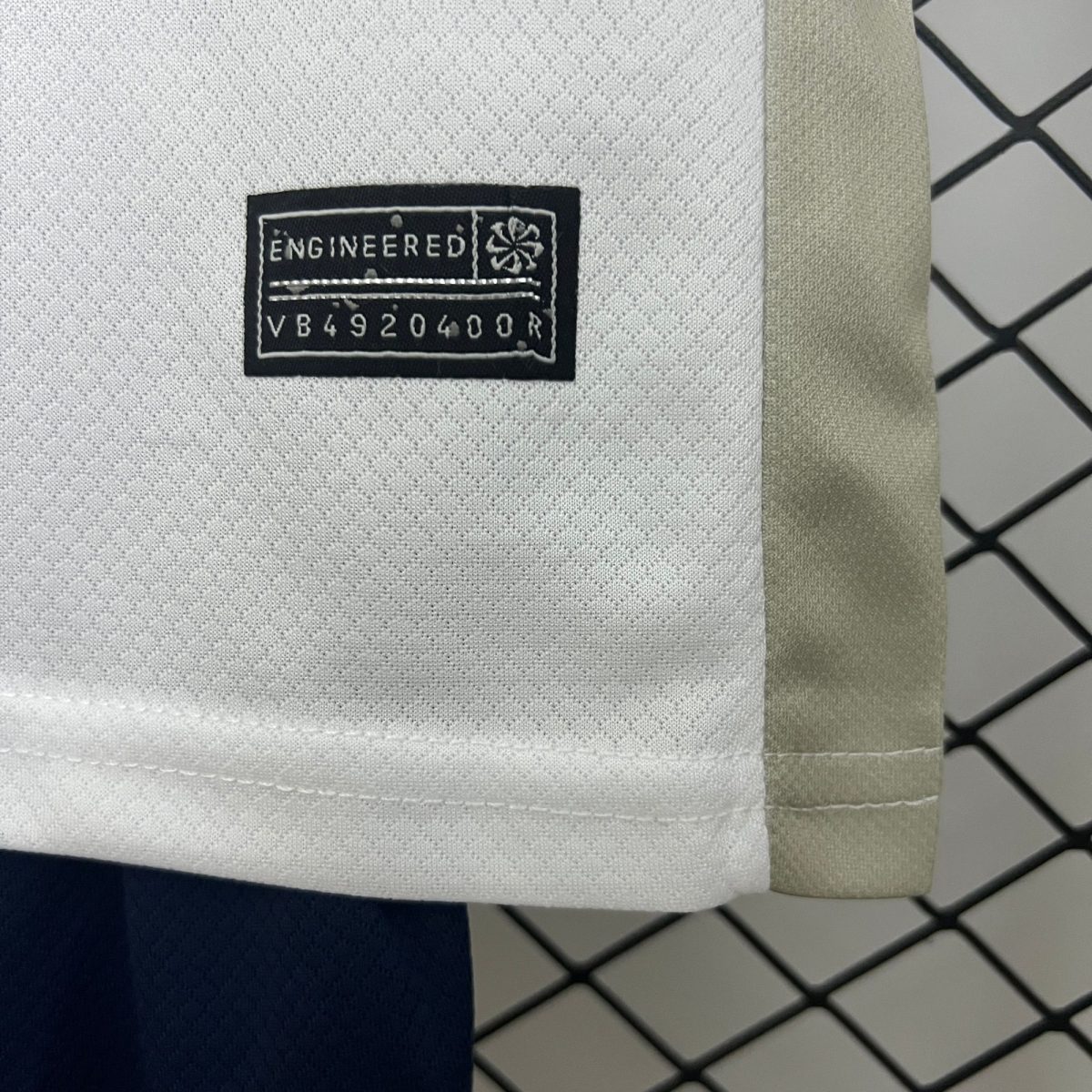 Close-up of a fabric swatch with a black label reading ENGINEERED and a code; light gray textured material with beige trim and a chain-link fence in the background.