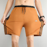 Man wearing orange athletic shorts with a black drawstring and black side stripes; logo on the left thigh.