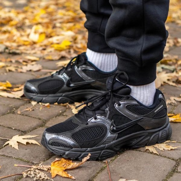 Close-up of black athletic sneakers with gray accents on a brick path, autumn leaves scattered around.