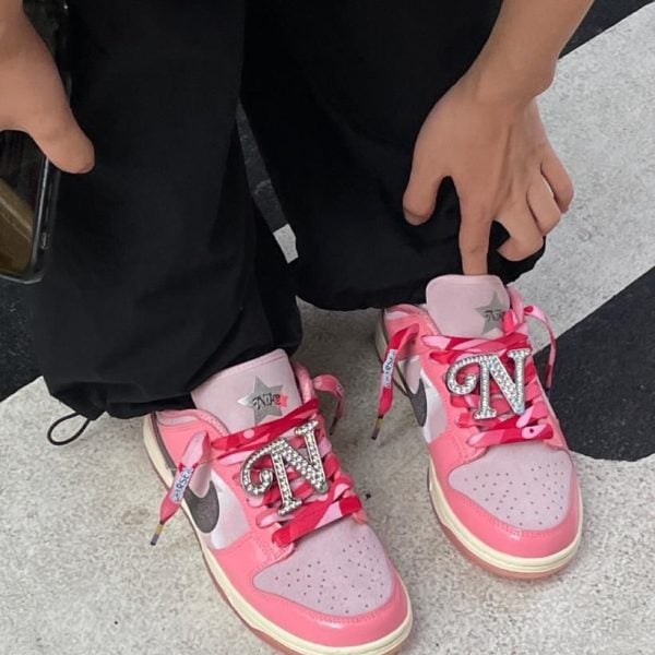 Close-up of pink and white sneakers with large rhinestone 'TN' charms on the laces worn by a person in black pants.
