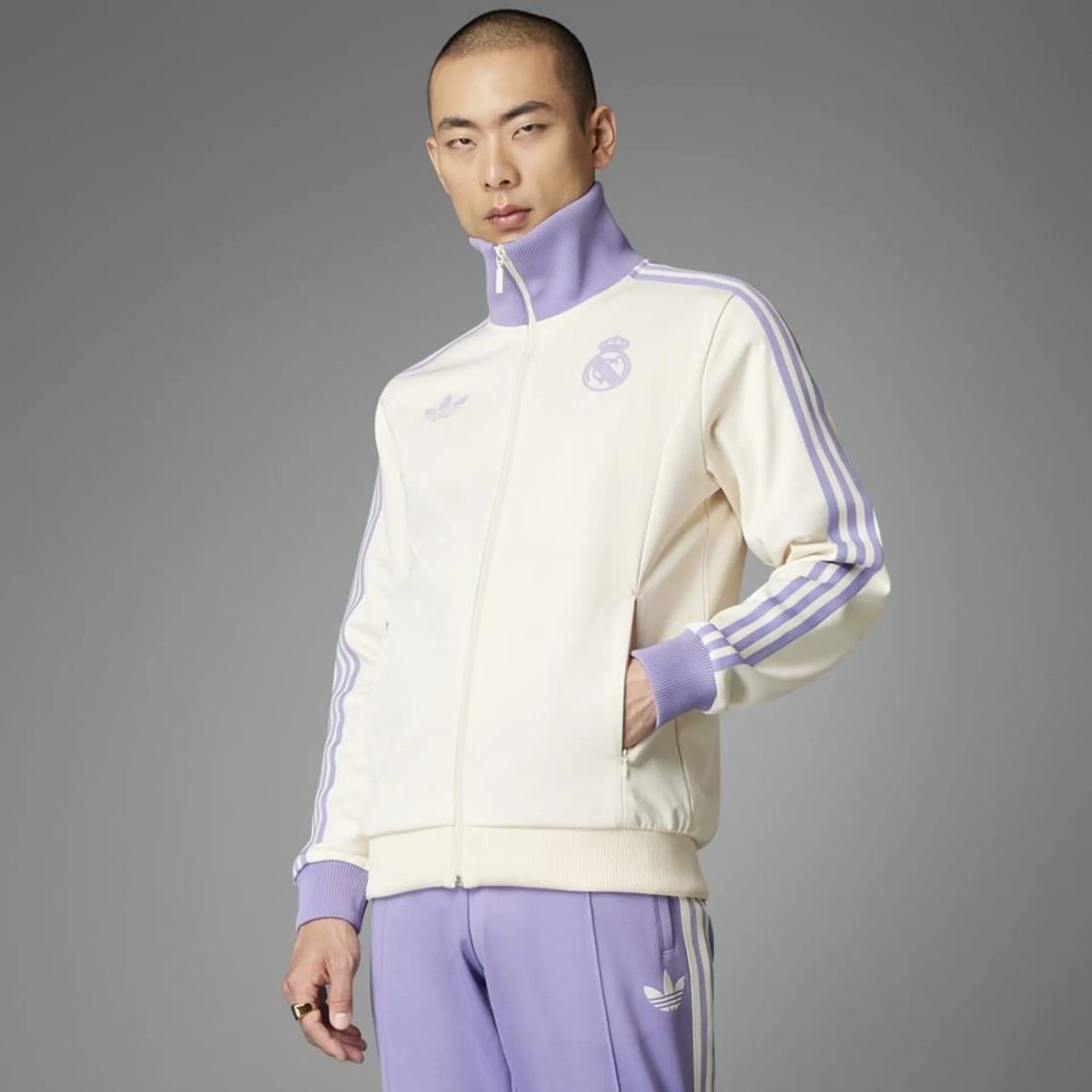 Man modeling a cream and lavender Adidas tracksuit with striped sleeves, standing against a gray studio backdrop.