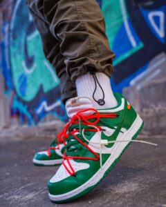 Close-up of green and white sneakers with red laces on a pavement, graffiti wall in the background.