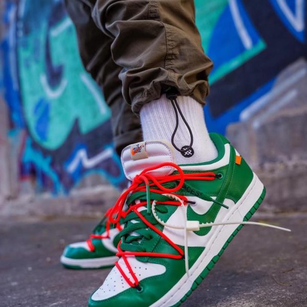 Close-up of green and white sneakers with red laces on a pavement, graffiti wall in the background.