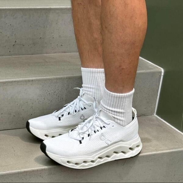 White athletic sneakers with a chunky wavy sole on gray concrete stairs, worn with white crew socks.