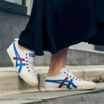 Pair of white low-top sneakers with blue and red stripes resting on a curb, worn with a dark skirt or dress.