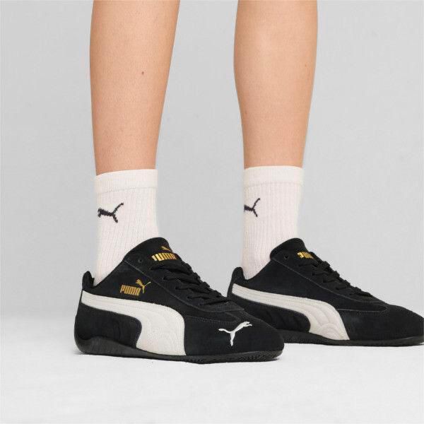 Pair of black Puma sneakers with white soles and gold branding, worn with white socks against a light gray background.