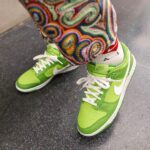 Bright lime-green Nike sneakers with white laces on a speckled gray floor, colorful patterned pants partially covering the ankles.
