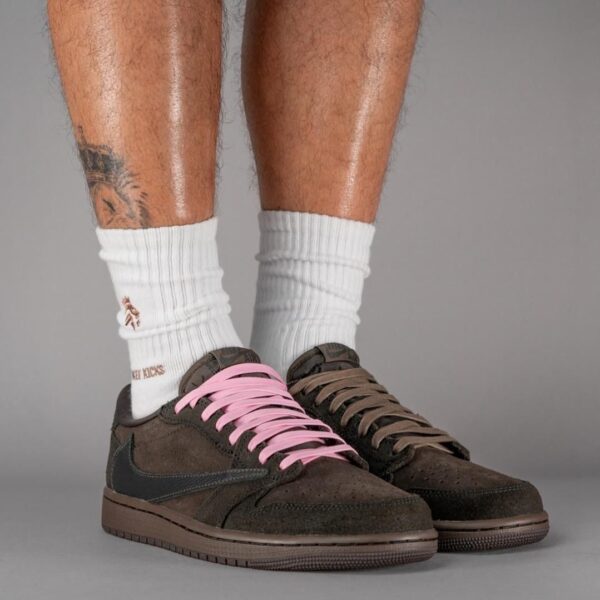 Brown suede sneakers with pink lace on the left shoe and brown lace on the right, worn with white crew socks and visible leg tattoos against a gray backdrop