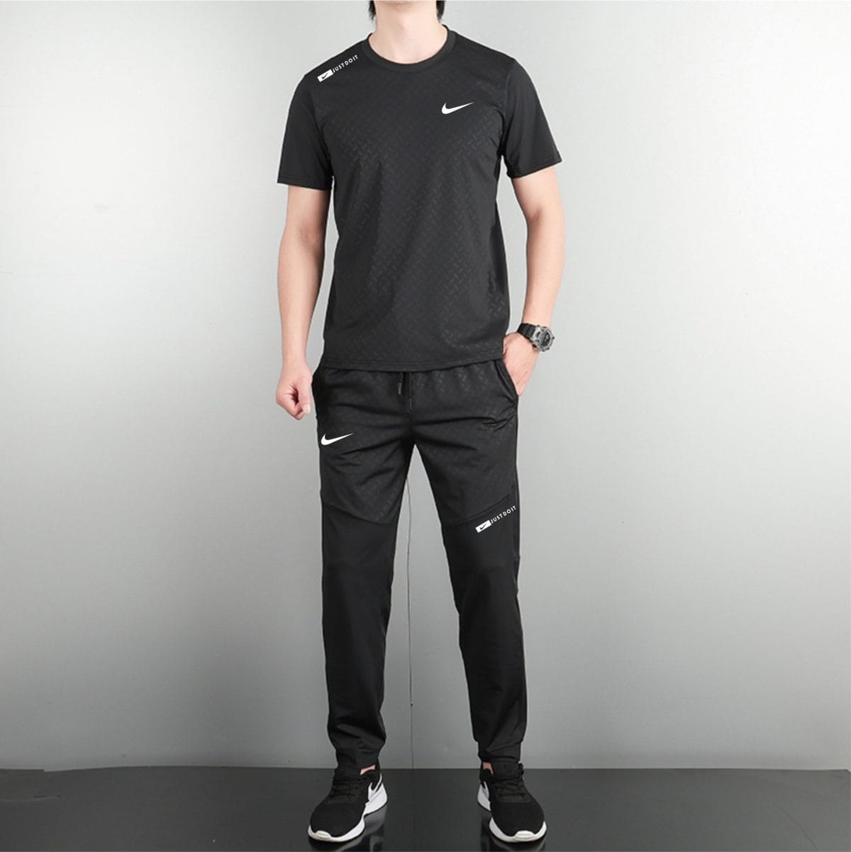 Full-body shot of a person wearing a black Nike athletic outfit, standing against a plain wall, hands at sides.