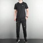 Full-body shot of a person wearing a black Nike athletic outfit, standing against a plain wall, hands at sides.