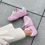 Pair of pink sneakers with white laces resting on a concrete sidewalk, worn with light cream ribbed pants.