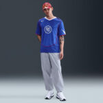 Young man in a blue sports jersey with number 90, gray baggy pants, white sneakers, and a red flame-pattern cap posing against a dark backdrop.