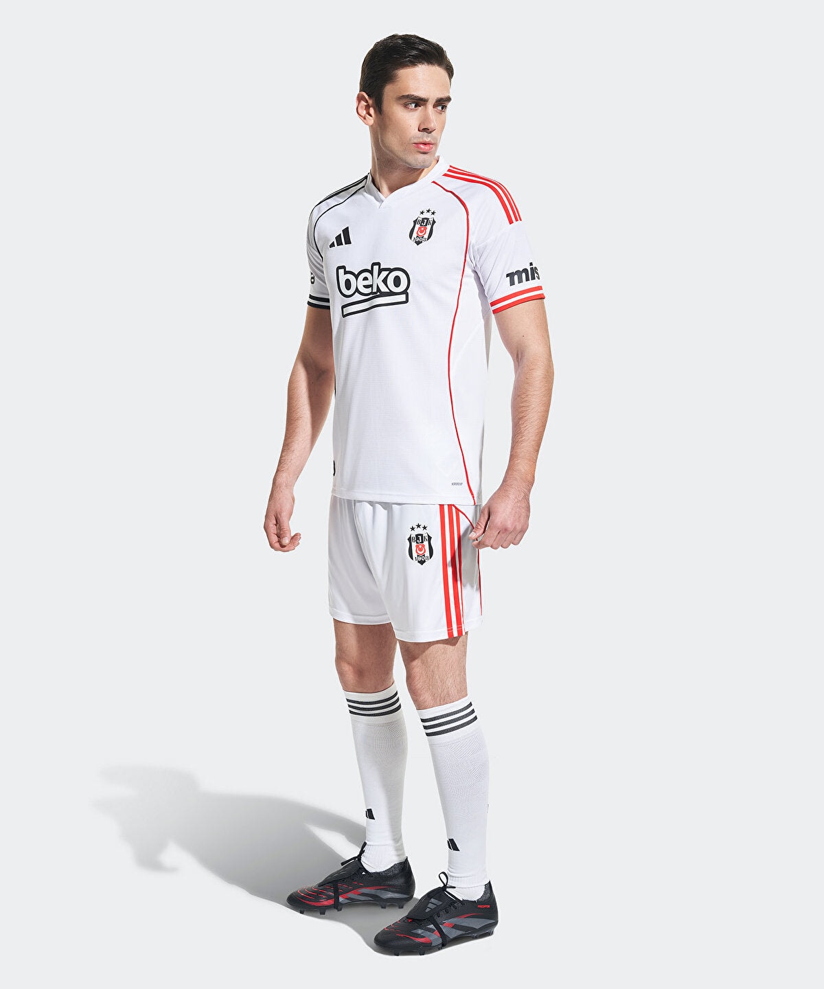 6KS4E0100001-01_3 | UAE SPORT STORE Male model in a white soccer jersey and shorts with red piping, black cleats, standing against a light background.