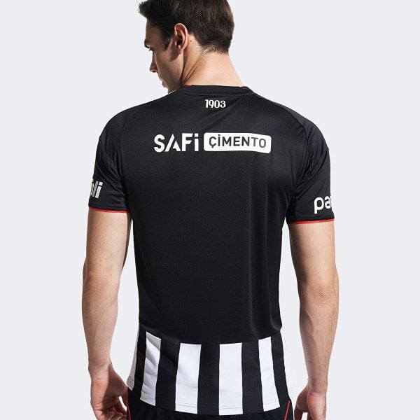 Back view of a man wearing a black soccer jersey with a white 'SAFi CIMENTO' logo on the upper back and black-and-white striped fabric at the waist.