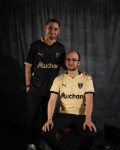 Two men pose for a portrait in matching soccer jerseys; one in black standing, the other in gold seated, both with 'Auchan' sponsor logos.