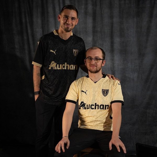 Two men pose for a portrait in matching soccer jerseys; one in black standing, the other in gold seated, both with 'Auchan' sponsor logos.