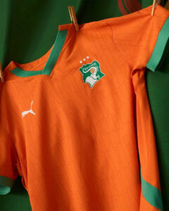 Orange football jersey with green trim and a Côte d'Ivoire crest on the chest, hanging with clothespins against a dark background, Puma logo visible.