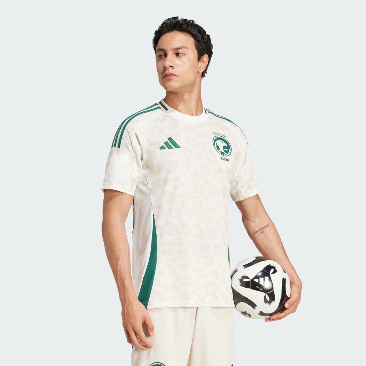 Male soccer player in a white Saudi Arabia jersey with green accents holding a soccer ball against a light gray background