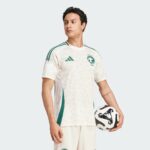 Male soccer player in a white Saudi Arabia jersey with green accents holding a soccer ball against a light gray background