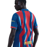 Back view of a dark-skinned man wearing a blue jersey with red diagonal stripes and a sponsor logo on the lower back.