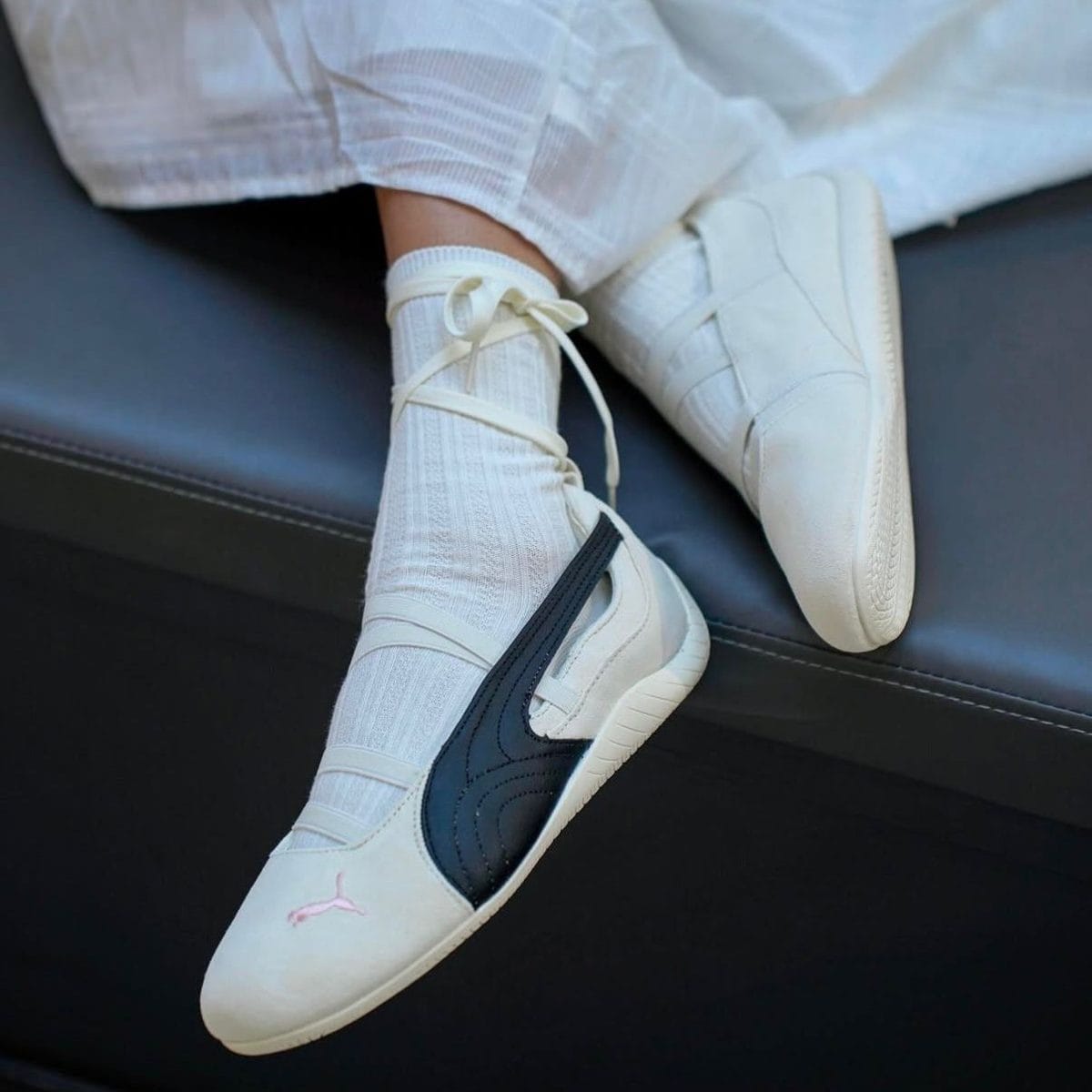 White sneakers with a dark blue swoosh and pink Jumpman emblem on the toe, worn with white socks and resting on a black couch.