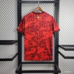 Back view of a red camouflage T-shirt on a wooden hanger, displayed on a black metal rack; gold text near the neckline reads 1921 2024.