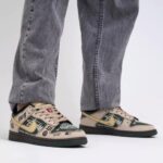 Low-top sneakers in beige and dark green with graffiti logos and a gold swoosh, paired with gray denim jeans.