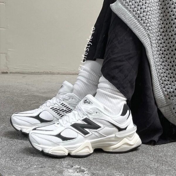 Pair of white chunky sneakers with black detailing and white crew socks resting on a concrete floor beside a grey knit garment.