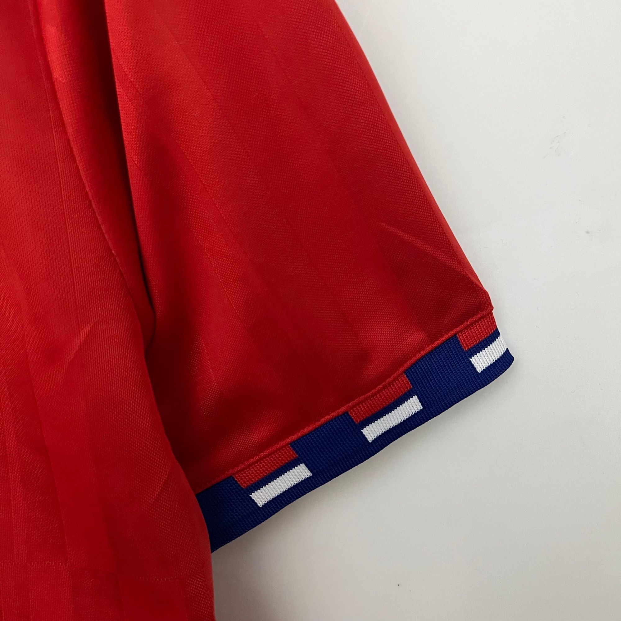 6b2037ca_6aa7a36f-54c5-49c1-aa01-4322a3dd67b3 | UAE SPORT STORE Close-up of a red garment sleeve with a blue, white-striped cuff edge and visible fabric texture.