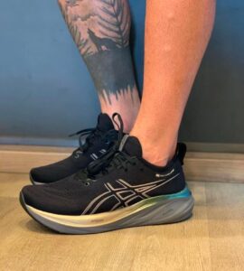 Black ASICS running shoes with a light beige sole on a wooden floor; visible tattooed lower leg with a forest/wolf design nearby.