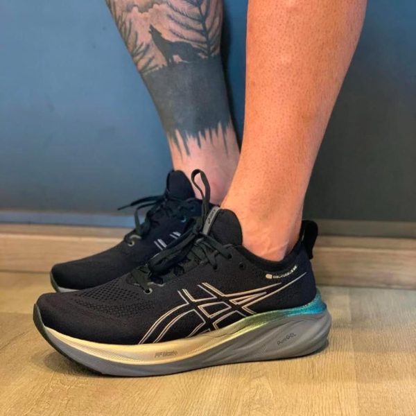 Black ASICS running shoes with a light beige sole on a wooden floor; visible tattooed lower leg with a forest/wolf design nearby.