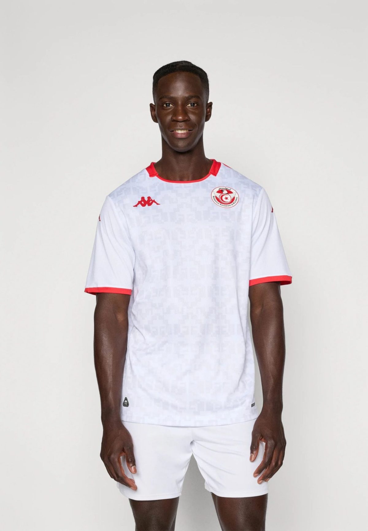 Portrait of a smiling man in a white soccer jersey with red trim, standing against a light background, crest visible on the chest.