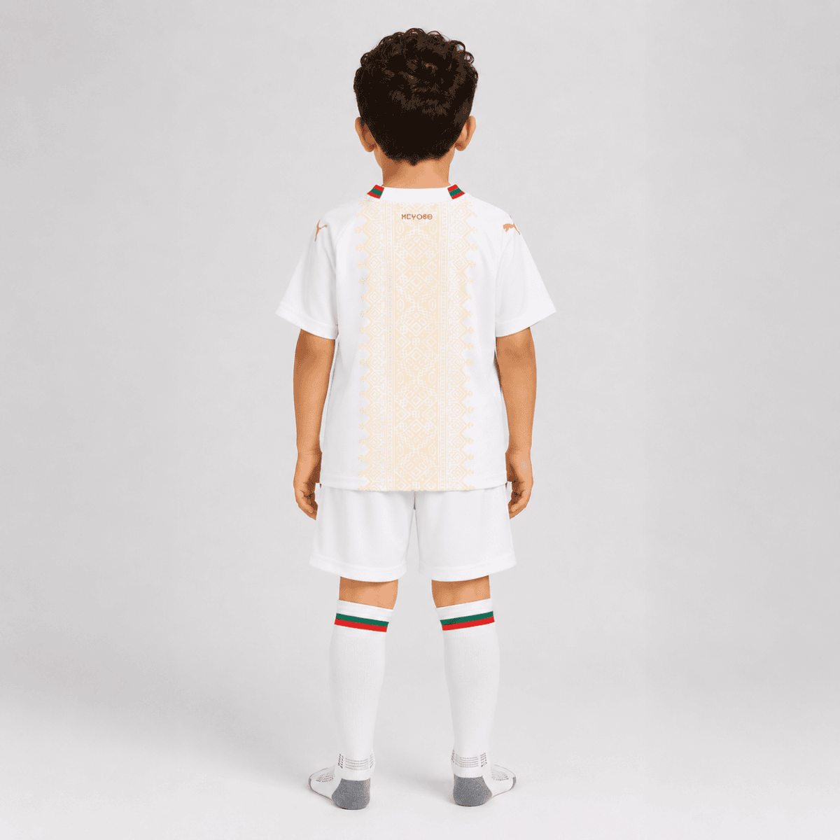Back view of a young boy wearing a white soccer kit with gold vertical embroidery, green-red trim on the collar, matching shorts and knee-high socks seen from behind.
