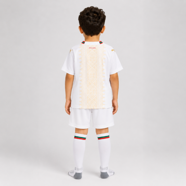 Back view of a young boy wearing a white soccer kit with gold vertical embroidery, green-red trim on the collar, matching shorts and knee-high socks seen from behind.