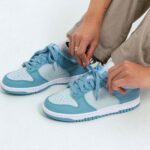 Person tying light blue and white sneakers on a white floor, close-up of hands threading laces.