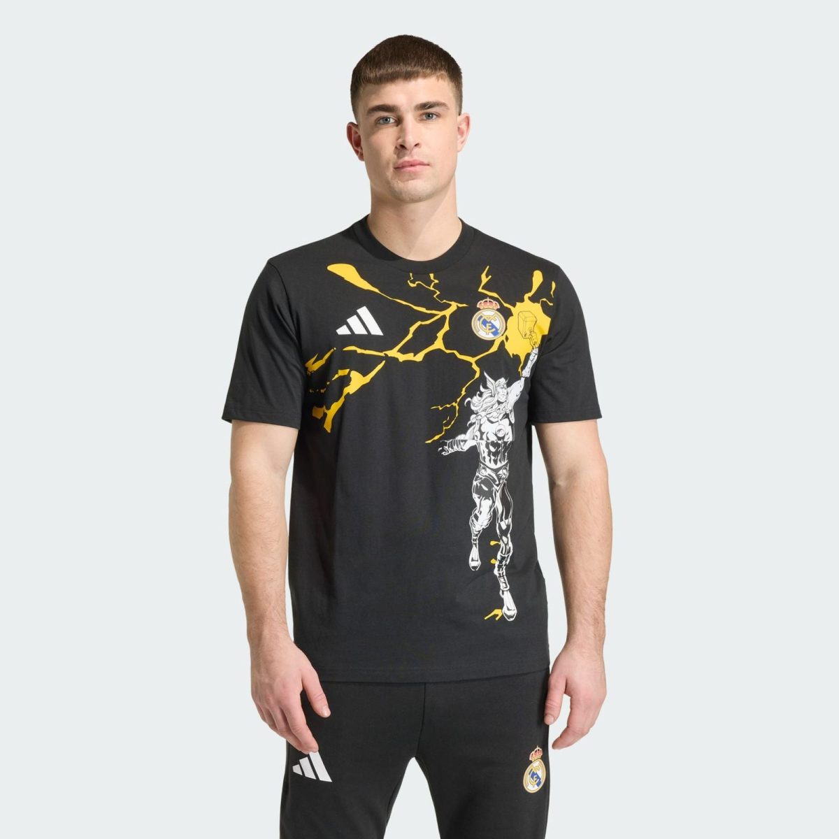 Man wearing a black Real Madrid graphic t-shirt with yellow lightning design and white Adidas logo, standing against a pale gray background.