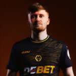 Portrait of a young man wearing a dark esports jersey with gold patterns and sponsor logos, standing against a brown background.