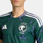 Close-up of a dark green Saudi football jersey with SAFF crest and Arabic text on the chest, white Adidas stripes on the shoulders.