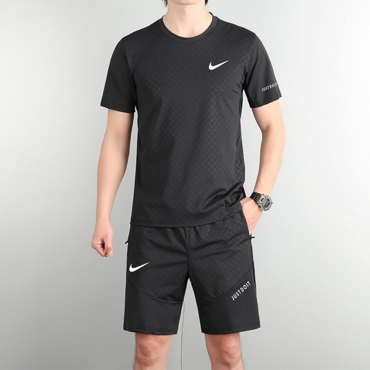 Person wearing a black Nike athletic shirt with a white swoosh logo, standing against a light grey wall with matching black shorts featuring logos.