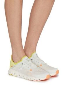 White On-running sneakers with yellow inner collar and orange-tipped soles worn on a person’s feet, side view.