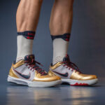 Pair of gold and gray Nike basketball shoes with maroon laces on a blue floor, worn with white athletic socks.