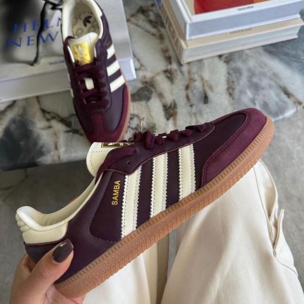 Maroon Adidas Samba sneaker with white stripes and gum sole, held in a hand; stack of books and a second shoe visible on a marble surface in the background.