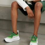 6df39dfd-e1d7-40ed-a12b-5808e9e3e2be | UAE SPORT STORE Person seated on a concrete surface, wearing a white blazer and green outfit, showcasing green-and-white athletic sneakers and holding a white bag.