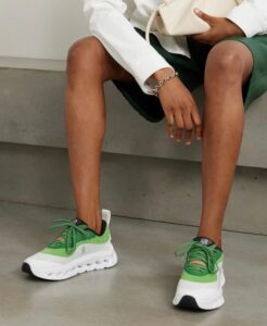 Person seated on a concrete surface, wearing a white blazer and green outfit, showcasing green-and-white athletic sneakers and holding a white bag.