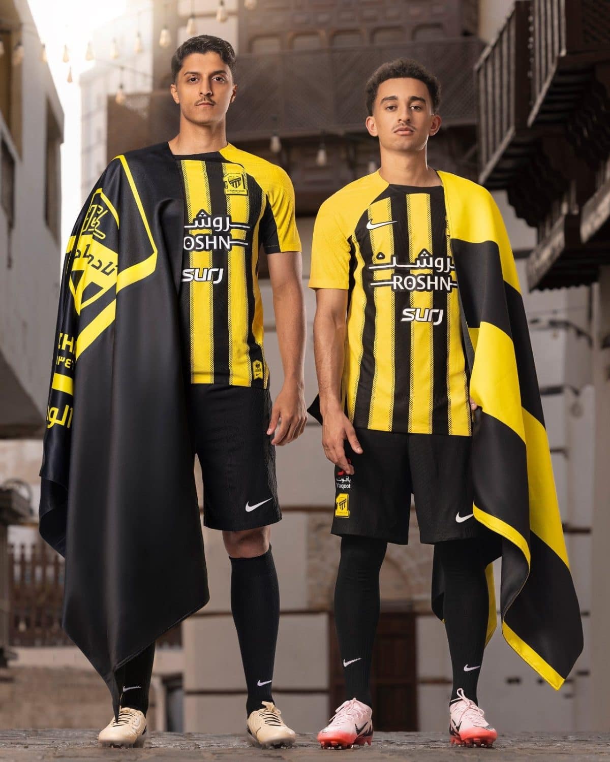 Two male soccer players model yellow and black striped jerseys with capes in an urban setting, Nike socks and cleats visible.
